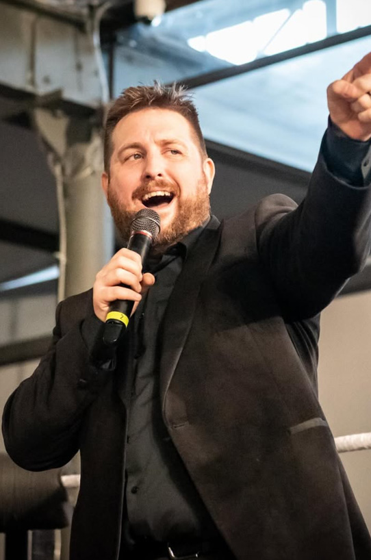 Tom Campbell, dressed in a black suit, energetically ring announcing with a microphone in hand, pointing towards the audience inside an indoor venue.